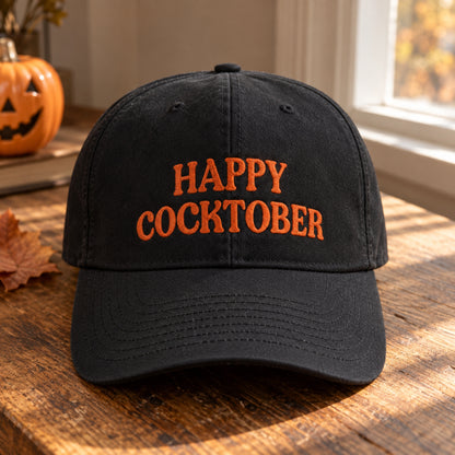 A black baseball cap with orange embroidered text reading 'HAPPY COCKTOBER' sits on a wooden table in a warm, autumnal setting with a jack-o'-lantern and leaf visible in the background.
