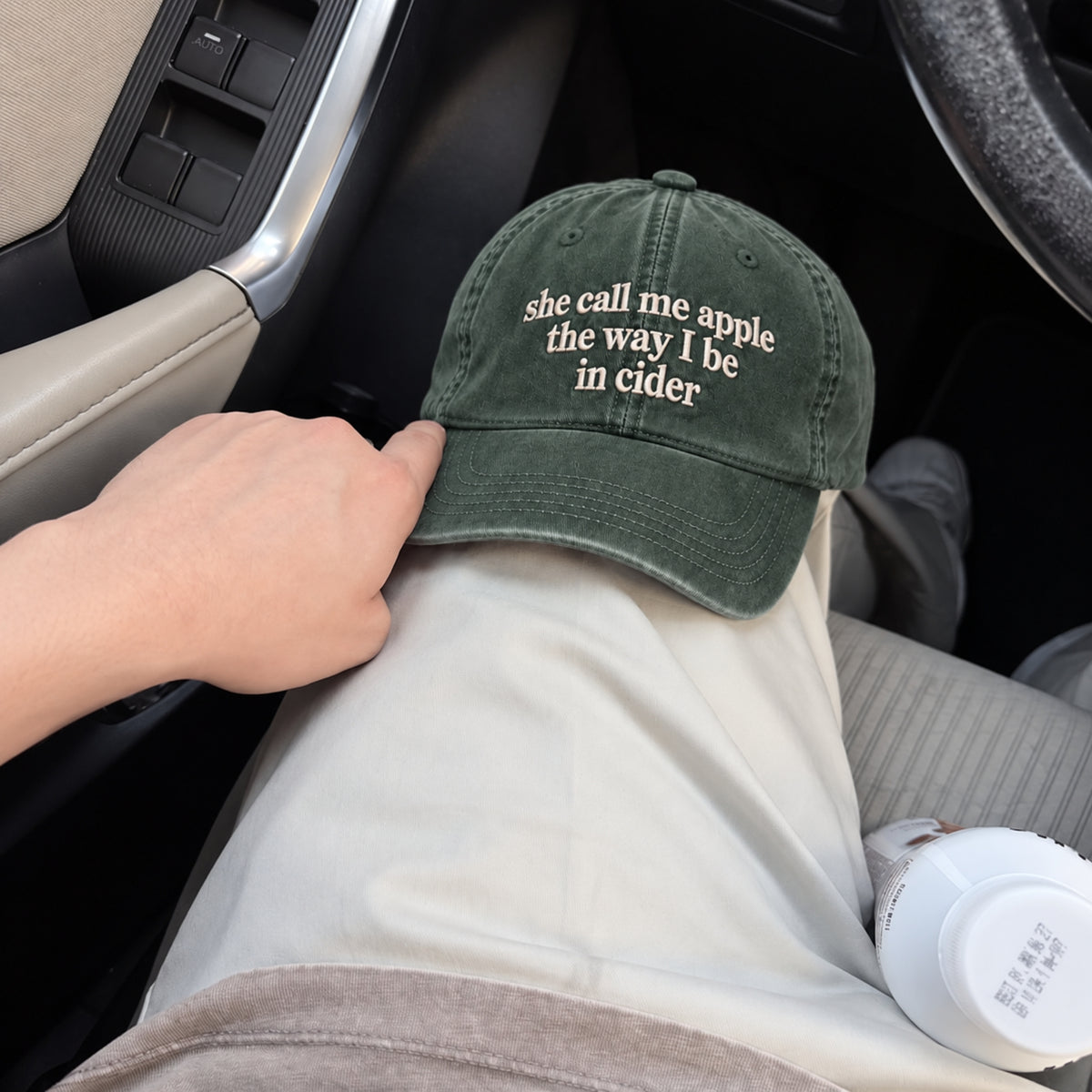 A person sitting in a car wearing an olive green baseball cap with the phrase 'she call me apple the way I be in cider' embroidered in white on the front. A hand is adjusting the cap, which is resting on their lap.
