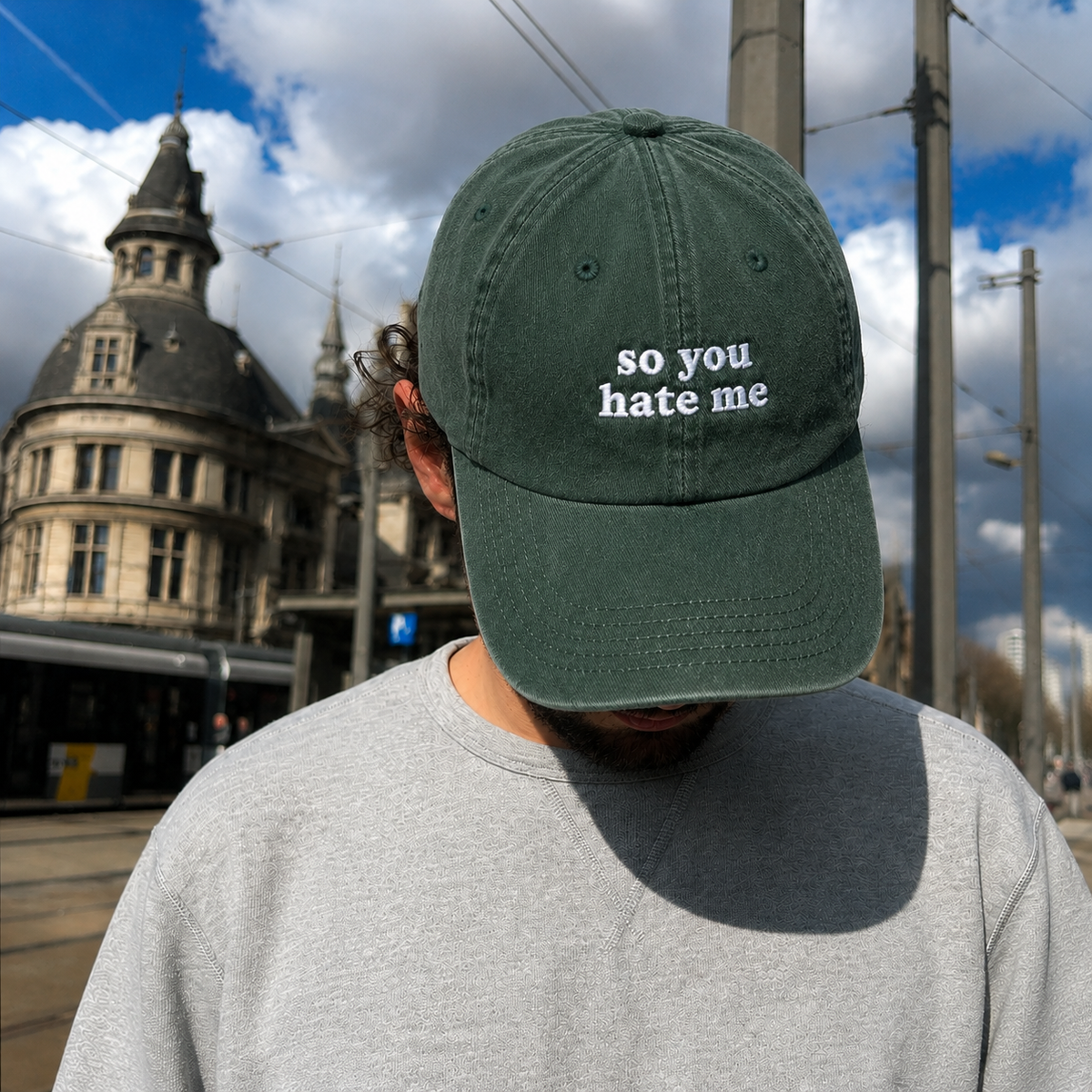 A man wearing a dark green baseball cap with the white embroidered text 'so you hate me' on the front, standing outdoors in front of a historic building and city street.