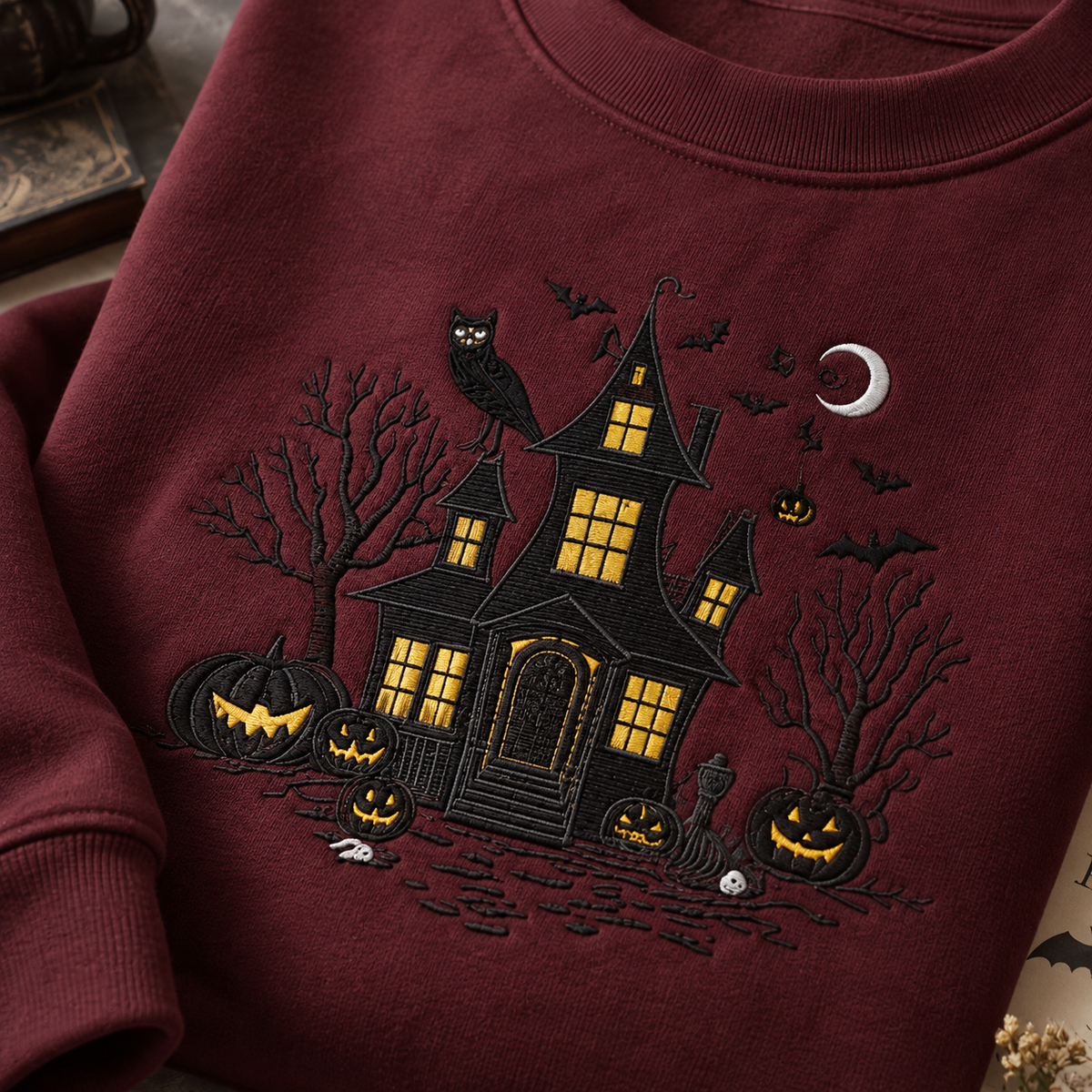 A close-up of a burgundy crewneck sweatshirt featuring a detailed black embroidered Halloween scene with a haunted house, jack-o'-lanterns, bats, an owl, and a crescent moon.