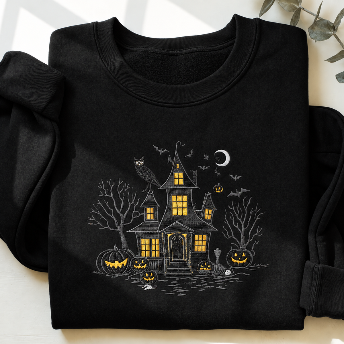 A black long-sleeve sweatshirt laid flat on a light surface, featuring a Halloween-themed house graphic with glowing yellow windows, bats, an owl, pumpkins, bare trees, and a crescent moon.