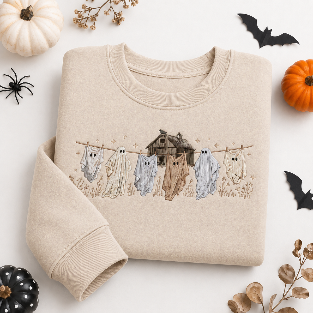 Sand beige embroidered Halloween sweatshirt featuring whimsical ghosts hanging on a clothesline with a rustic haunted house design, styled on a flat lay with autumn pumpkins and bats.
