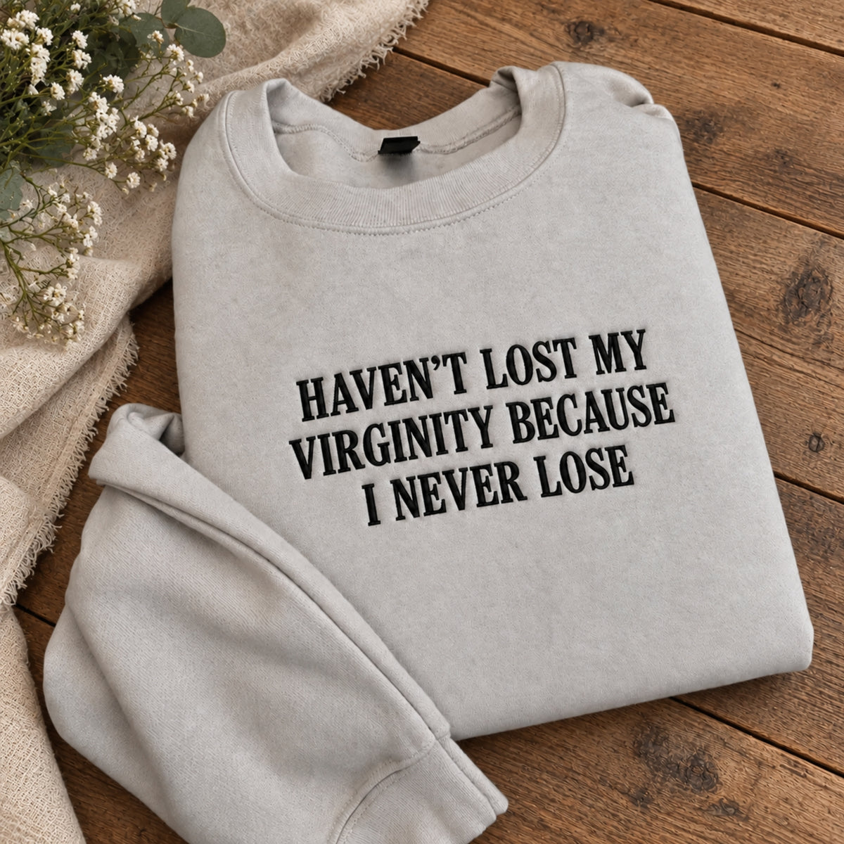 A light heather gray crewneck sweatshirt with black text reading 'HAVEN'T LOST MY VIRGINITY BECAUSE I NEVER LOSE', folded on a rustic wooden surface with a beige fabric and small white flowers as props.