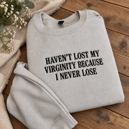 A light heather gray crewneck sweatshirt with black text reading 'HAVEN'T LOST MY VIRGINITY BECAUSE I NEVER LOSE', folded on a rustic wooden surface with a beige fabric and small white flowers as props.