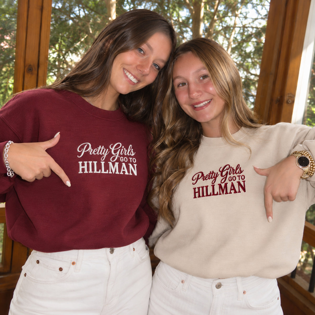 Two women posing together on a porch, each wearing a sweatshirt with the text 'Pretty Girls GO TO HILLMAN' printed on the chest. The woman on the left wears a burgundy sweatshirt, and the woman on the right wears a light beige sweatshirt.