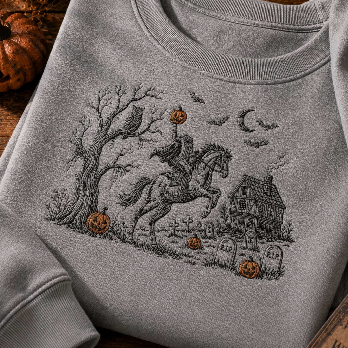 A light gray crewneck sweatshirt featuring a detailed black and white graphic of a rider on a galloping horse in a spooky graveyard, decorated with orange jack-o'-lanterns, under a crescent moon and bats.