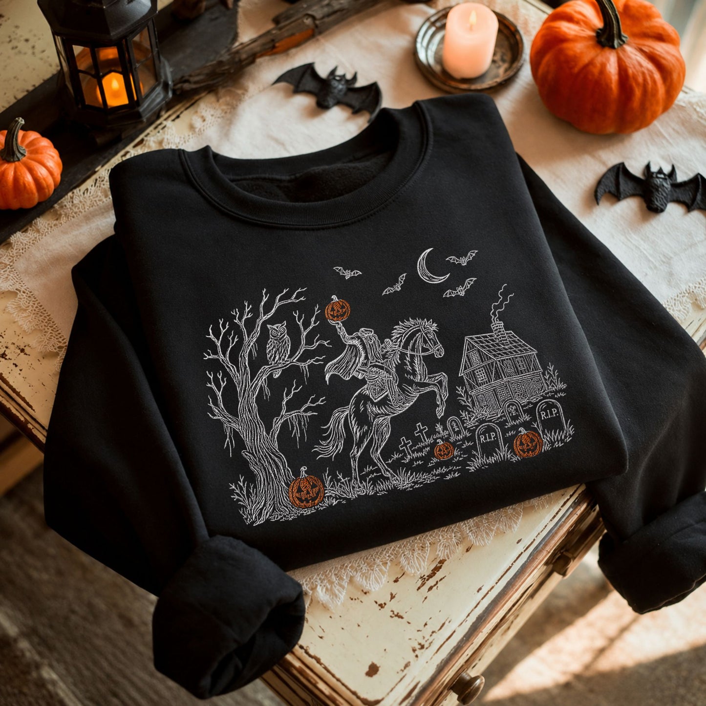 A black long-sleeve crewneck sweatshirt with a detailed white line-art graphic of a horse riding a ghostly figure in front of a haunted house, surrounded by pumpkins, bats, and a candle in a rustic Halloween-themed setting.