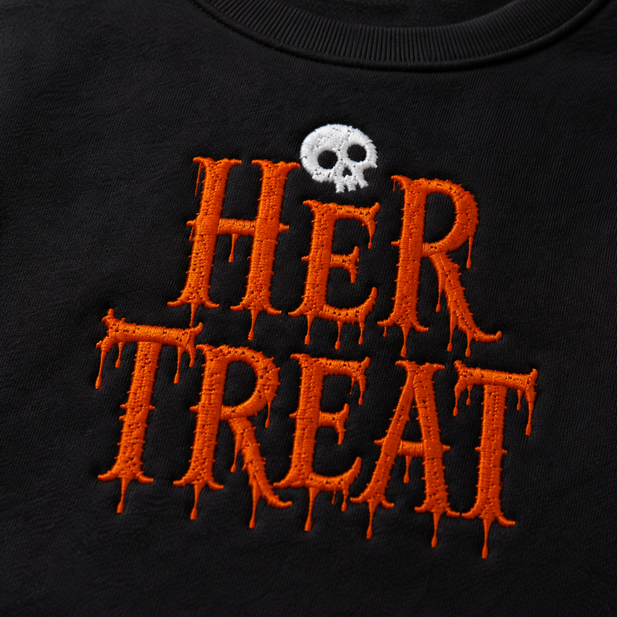 Close-up of a black garment featuring large orange embroidered dripping text reading 'HER TREAT' with a small white skull above the lettering.