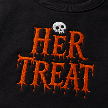 Close-up of a black garment featuring large orange embroidered dripping text reading 'HER TREAT' with a small white skull above the lettering.