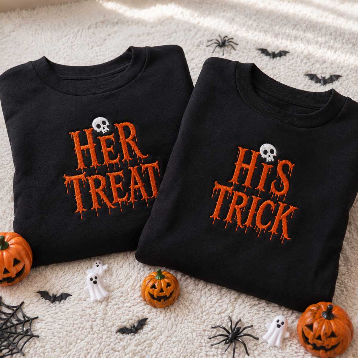 Two black long-sleeve T-shirts laid flat on a cream textured blanket with Halloween props. One shirt reads "HER TREAT" and the other reads "HIS TRICK" in orange dripping lettering, each with a small white skull above the text.