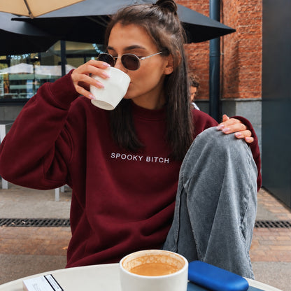 A woman wearing a burgundy sweatshirt with the text 'SPOOKY BITCH' in white, sitting at an outdoor cafe table and drinking from a white mug.