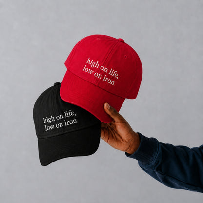 Two baseball caps are shown against a light gray background: one red cap held by a hand and one black cap behind it. Both caps have the phrase 'high on life, low on iron' embroidered in white on the front.