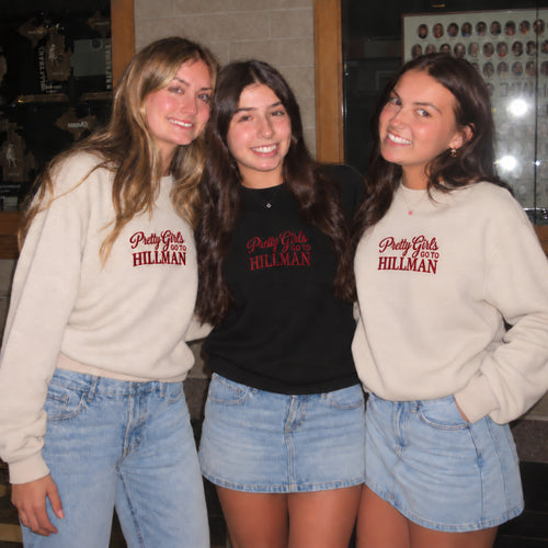 Three young women posing together indoors, wearing 'Pretty Girls Only HILLMAN' sweatshirts and light blue denim bottoms. The woman on the left wears a cream sweatshirt, the woman in the center wears a black sweatshirt, and the woman on the right wears a cream sweatshirt.