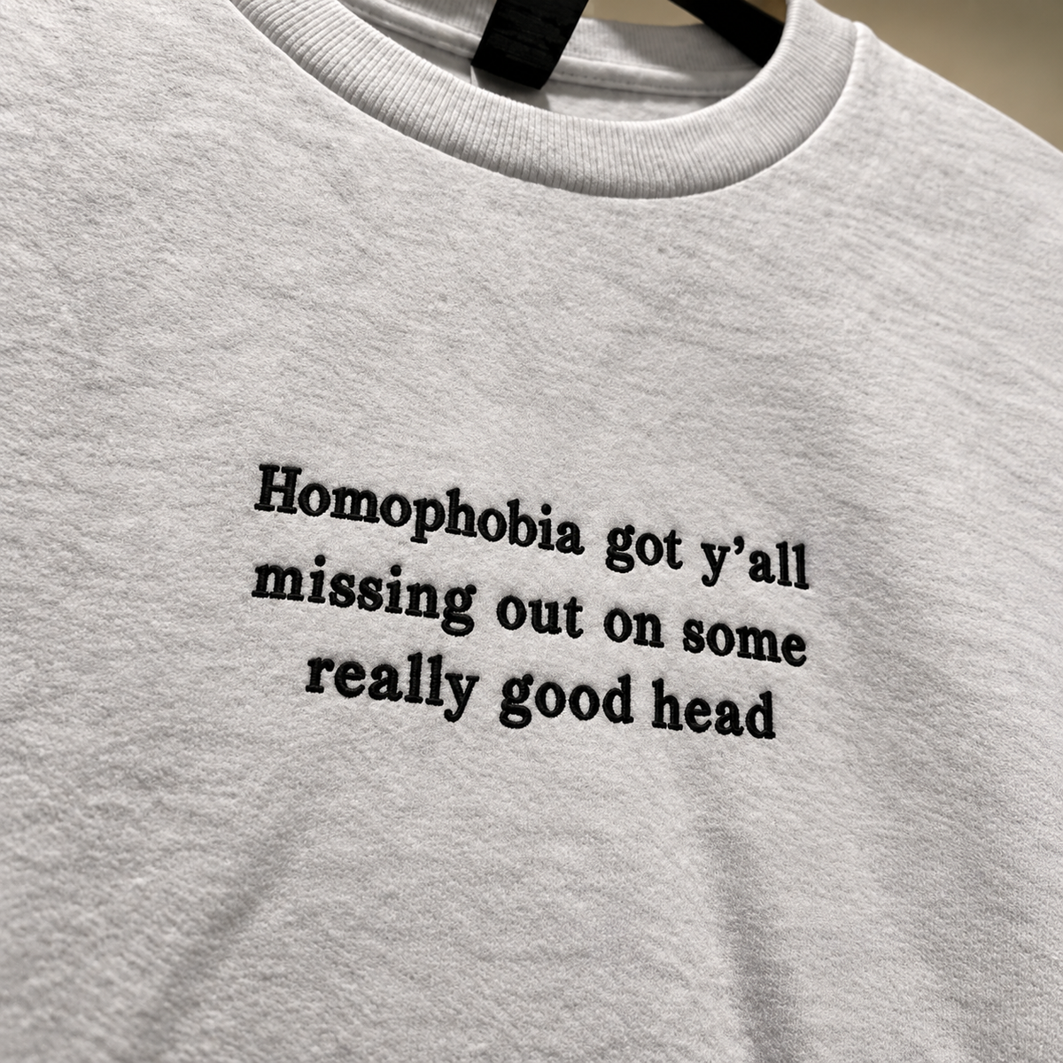 A close-up of a light gray heather crewneck t-shirt hanging on a black hanger, featuring the black printed text 'Homophobia got y'all missing out on some really good head'.