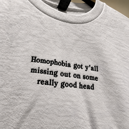 A close-up of a light gray heather crewneck t-shirt hanging on a black hanger, featuring the black printed text 'Homophobia got y'all missing out on some really good head'.