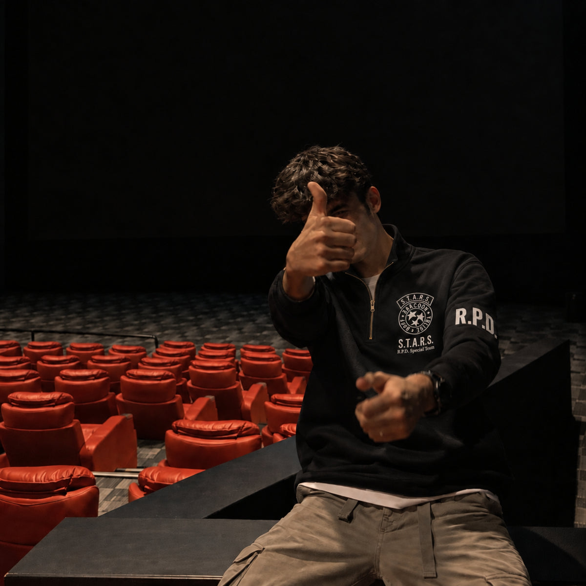 Man wearing a black quarter-zip sweatshirt with embroidered S.T.A.R.S. Raccoon Police Department chest emblem and R.P.D. sleeve text, seated in a dark theater with red seats.
