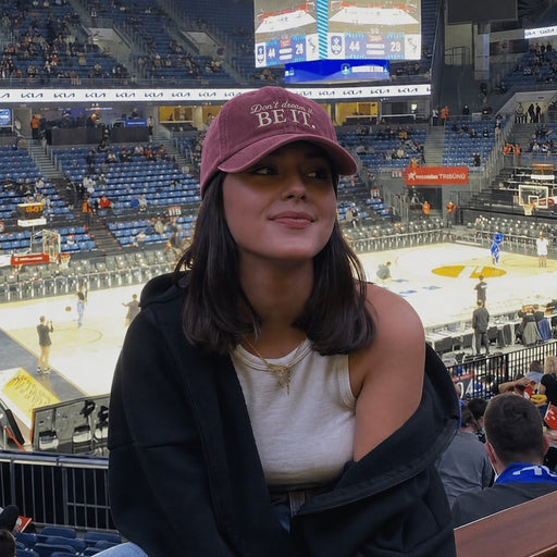 A woman in a crowded indoor arena wearing a maroon baseball cap with 'Dad, always BE IT.' embroidered on the front, a black cardigan, and a white tank top, with the basketball court and stadium seating in the background.