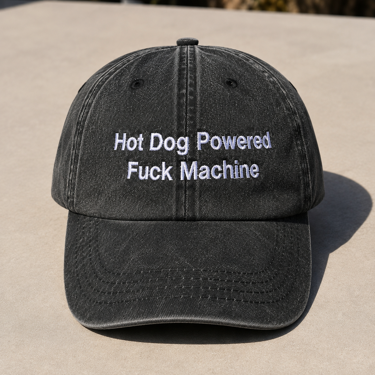 A washed black baseball cap with white embroidered text reading 'Hot Dog Powered Fuck Machine' on the front, photographed outdoors on a light concrete surface.