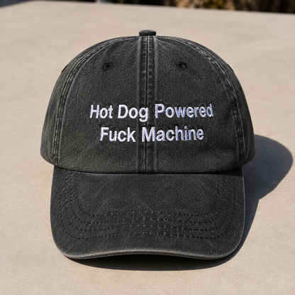 A washed black baseball cap with white embroidered text reading 'Hot Dog Powered Fuck Machine' on the front, photographed outdoors on a light concrete surface.