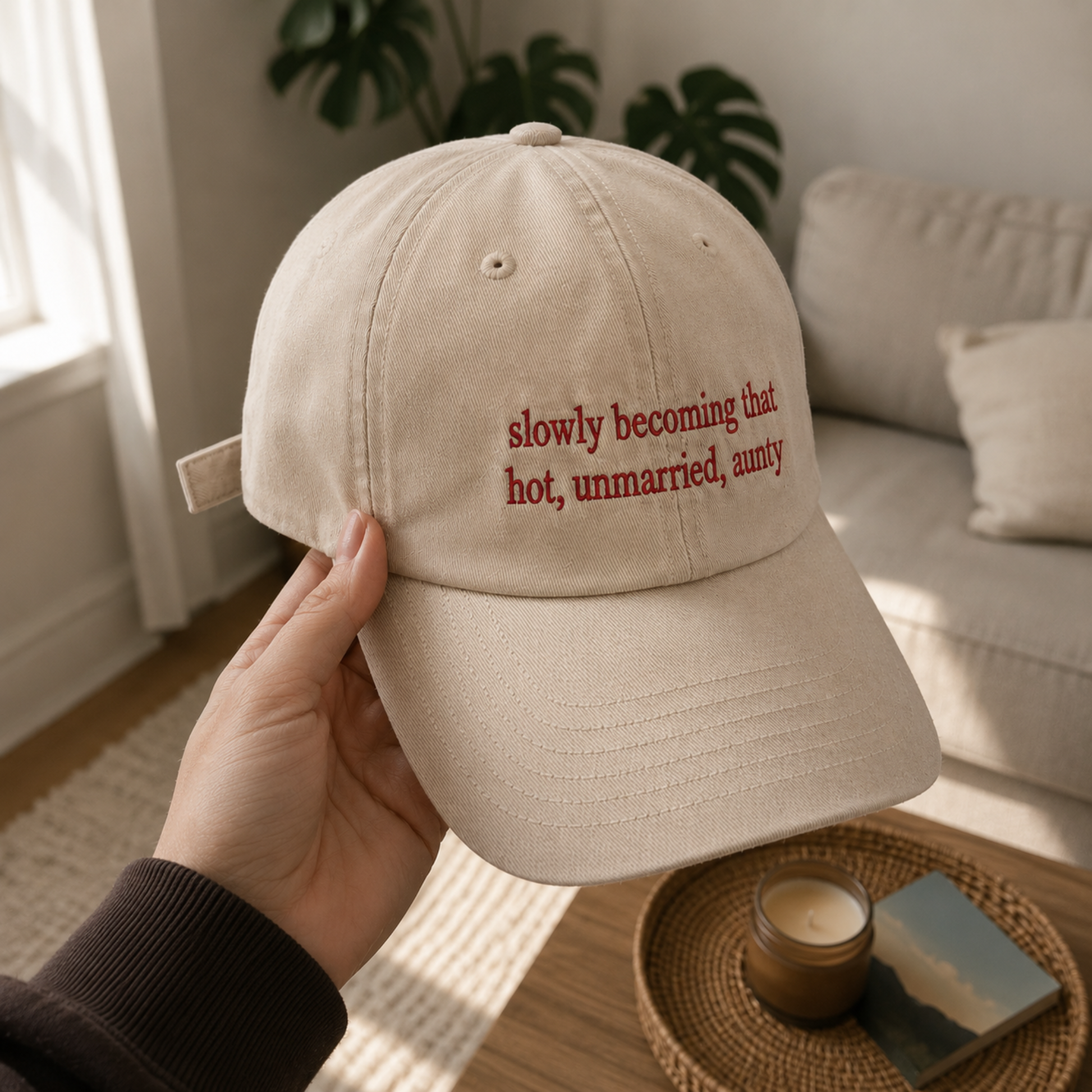 A hand holds a beige baseball cap with red embroidered text reading 'slowly becoming that hot, unmarried, aunty' in a cozy living room setting.