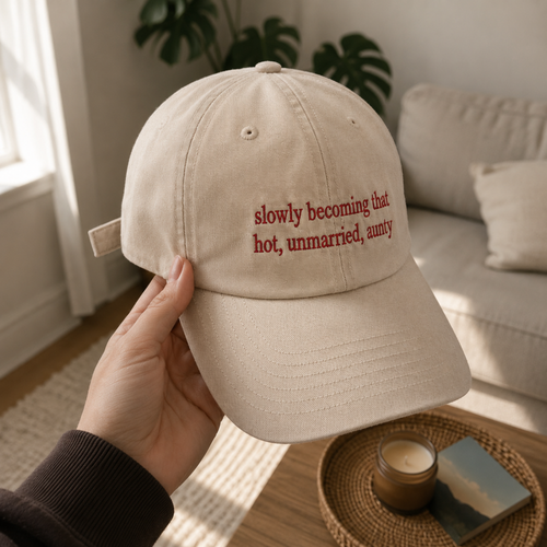 A hand holds a beige baseball cap with red embroidered text reading 'slowly becoming that hot, unmarried, aunty' in a cozy living room setting.