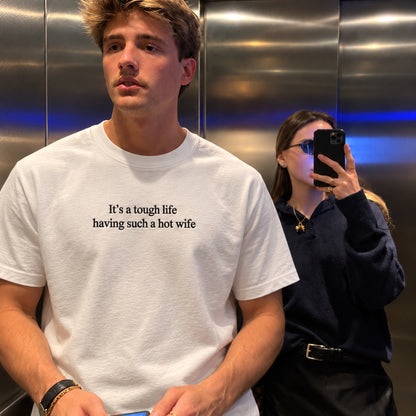 A man wearing a white crewneck t-shirt with the black printed text 'It's a tough life having such a hot wife' stands in front of a mirror inside an elevator, with a woman taking a selfie in the background.