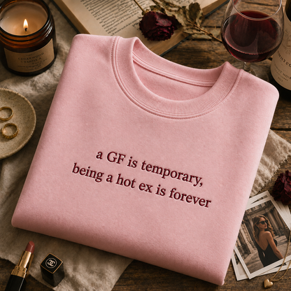 Light pink crewneck sweatshirt neatly folded on a wooden table next to a lit candle, lipstick, and a glass of red wine, featuring embroidered text that reads "a GF is temporary, being a hot ex is forever".