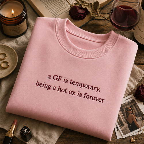 Light pink crewneck sweatshirt neatly folded on a wooden table next to a lit candle, lipstick, and a glass of red wine, featuring embroidered text that reads "a GF is temporary, being a hot ex is forever".