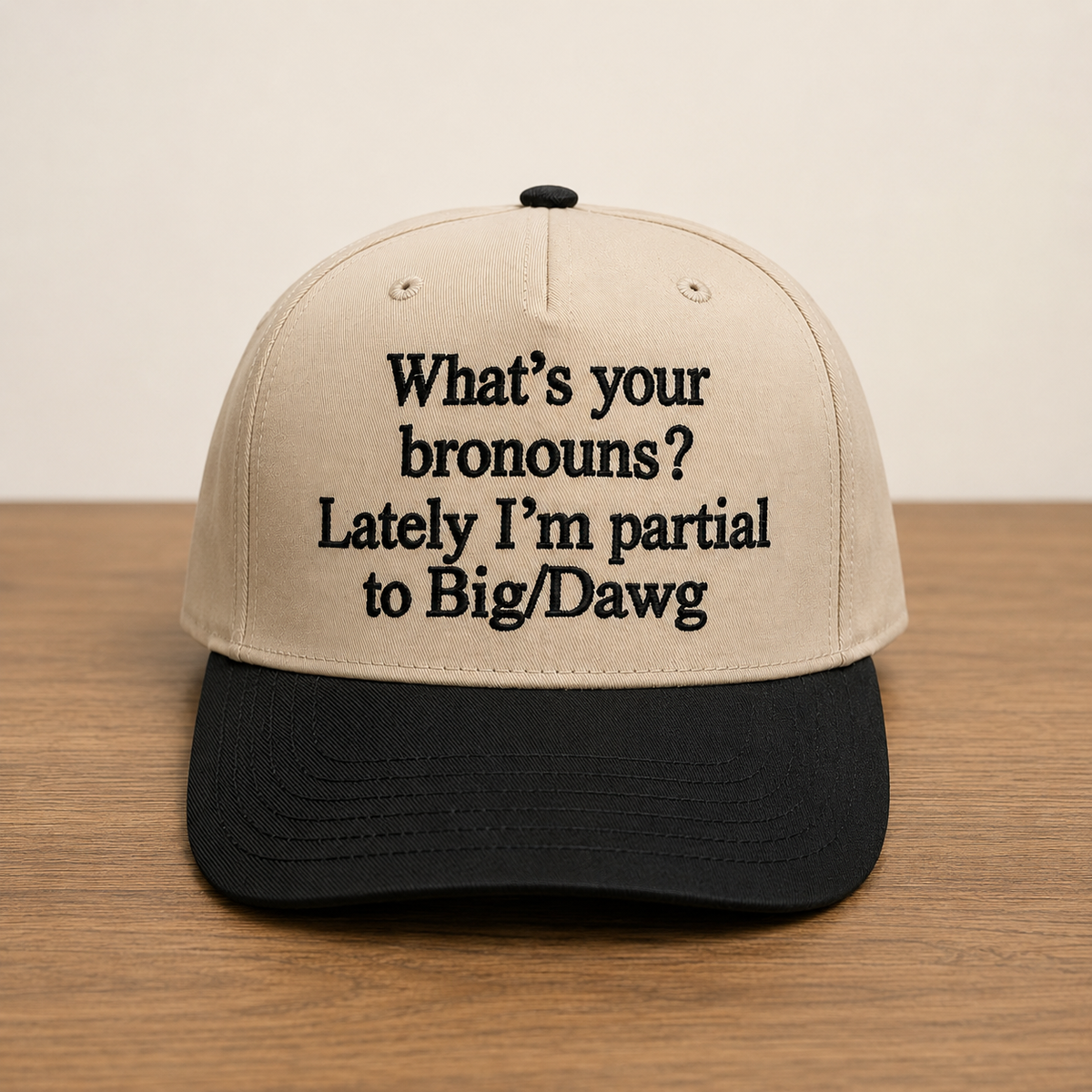 A beige and black baseball cap with black embroidered text reading 'What's your bronouns? Lately I'm partial to Big/Dawg', displayed on a wooden surface against a light beige background.