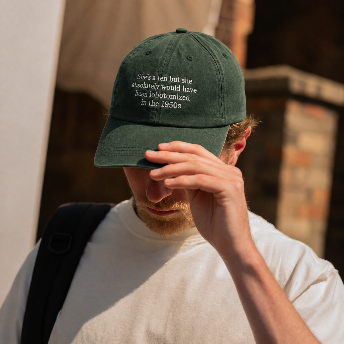 A man wearing a dark green baseball cap with white embroidered text that reads 'She's a ten but she absolutely would have been lobotomized in the 1850s'. He is wearing a white t-shirt and a black backpack strap, with a blurred outdoor background.