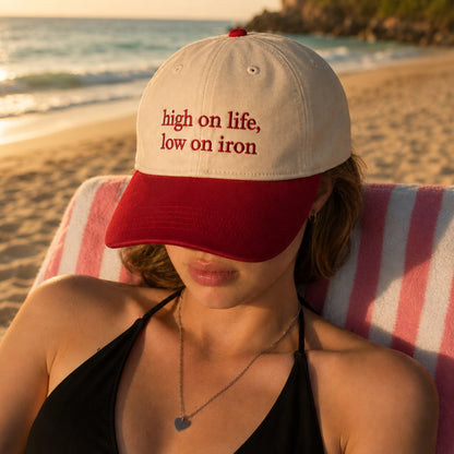 A woman sitting on a pink and white striped beach chair on a sandy beach, wearing a cream and red baseball cap with the embroidered text 'high on life, low on iron'.