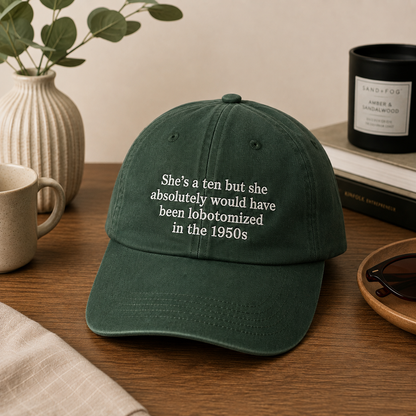 A dark green baseball cap with white embroidered text reading 'She's a ten but she absolutely would have been lobotomized in the 1950s' sits on a wooden table in a styled home setting with a mug, plant, sunglasses, and books.