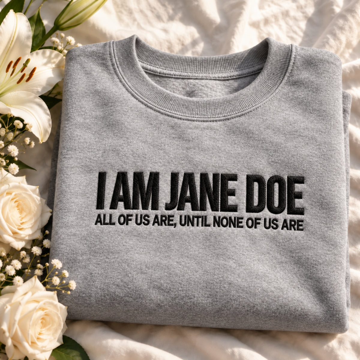 A folded light gray crewneck sweatshirt with black text reading 'I AM JANE DOE' and 'ALL OF US ARE, UNTIL NONE OF US ARE' lies on a cream fabric surface next to white flowers.