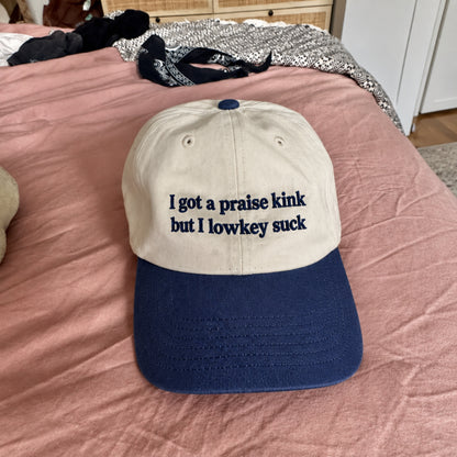 A cream and navy embroidered baseball cap featuring the phrase “I got a praise kink but I lowkey suck,” photographed on a pink bedspread, with clothing visible in the background.