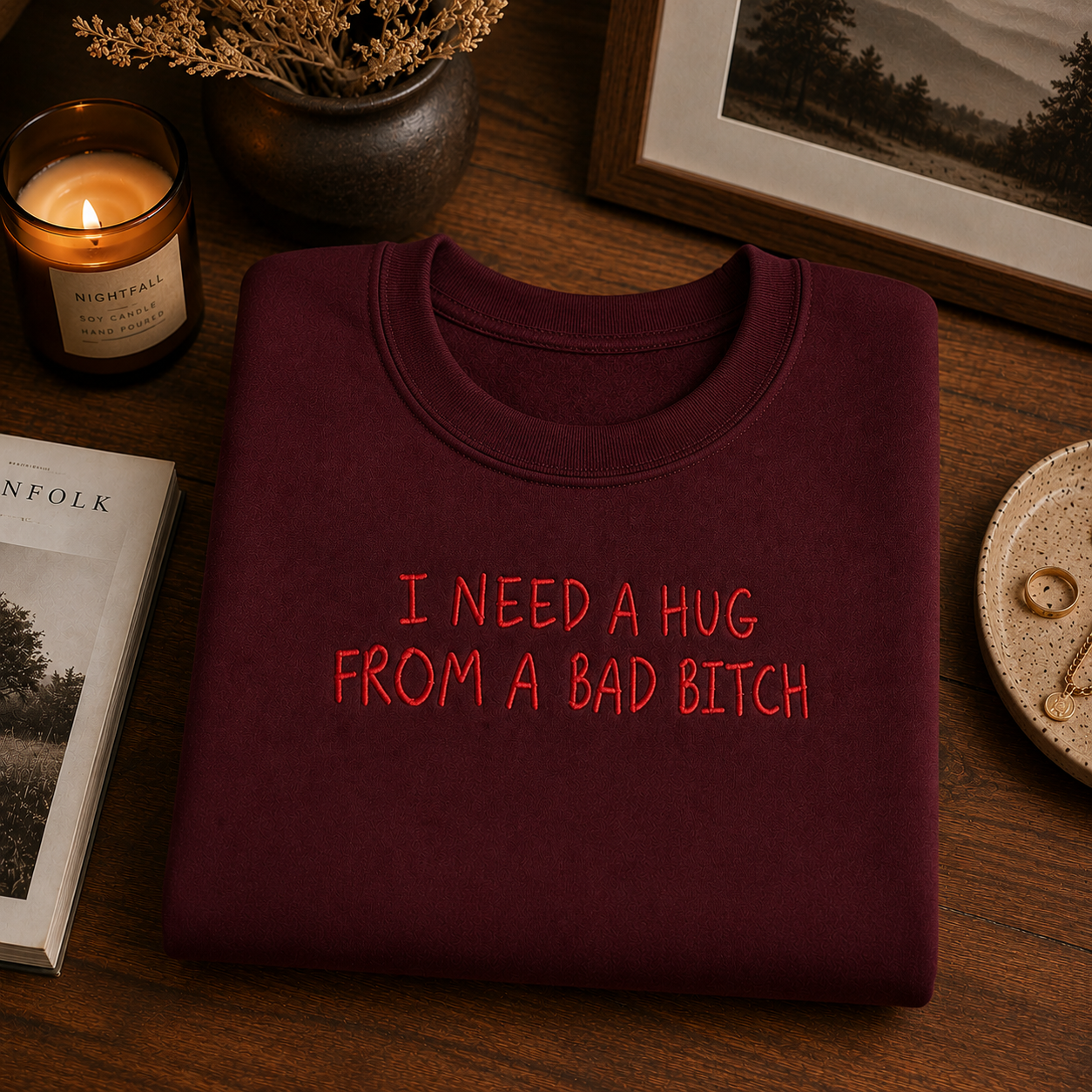 A folded burgundy crewneck sweatshirt with red embroidered text reading 'I NEED A HUG FROM A BAD BITCH' is displayed on a wooden table surrounded by cozy lifestyle props including a lit candle, a book, and a framed picture.