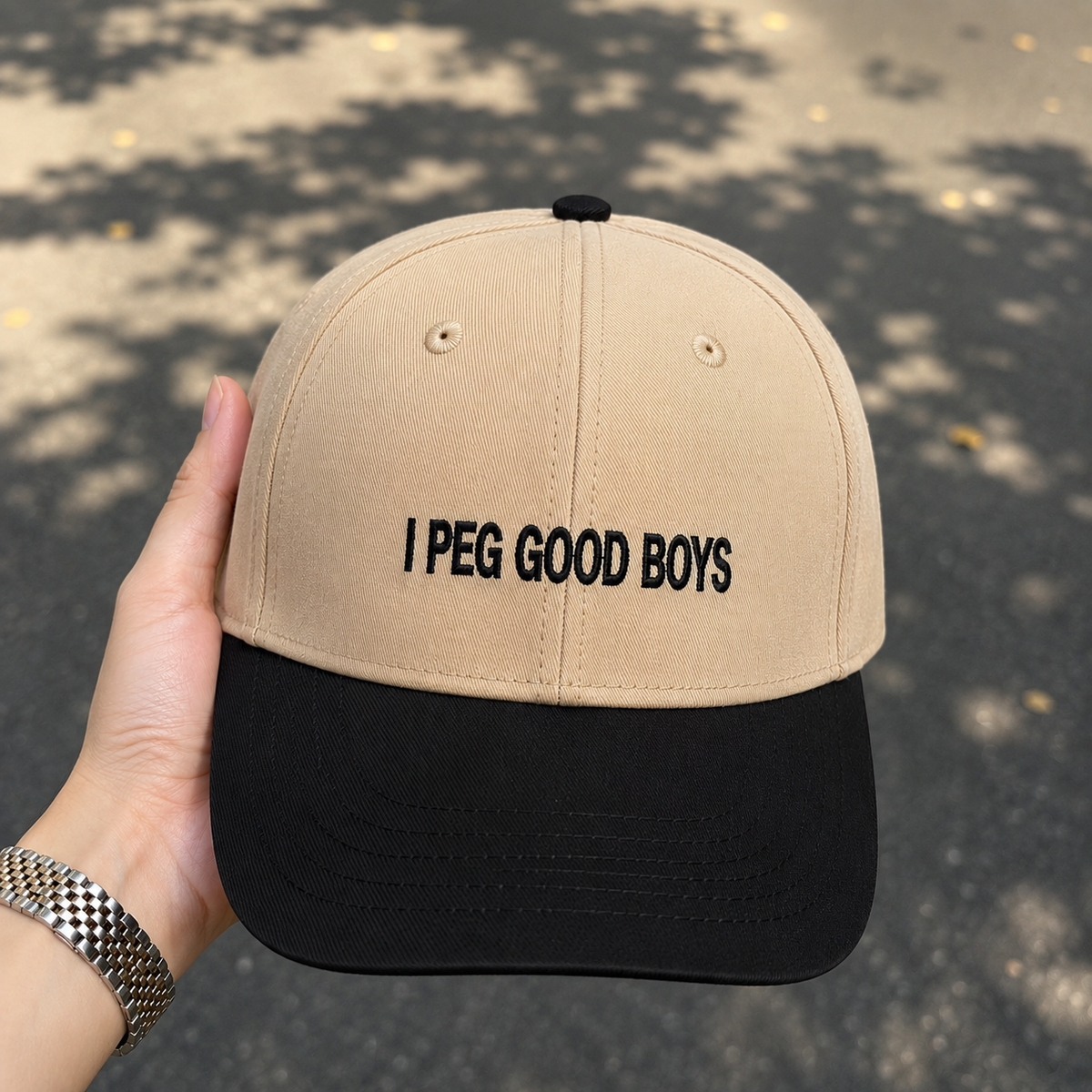 A beige and black baseball cap held by a hand, featuring the embroidered text 'I PEG GOOD BOYS' on the front. The cap has a beige crown and a black visor, set against a blurred outdoor background with tree shadows.