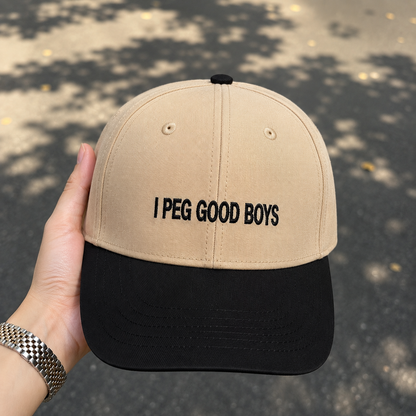 A beige and black baseball cap held by a hand, featuring the embroidered text 'I PEG GOOD BOYS' on the front. The cap has a beige crown and a black visor, set against a blurred outdoor background with tree shadows.