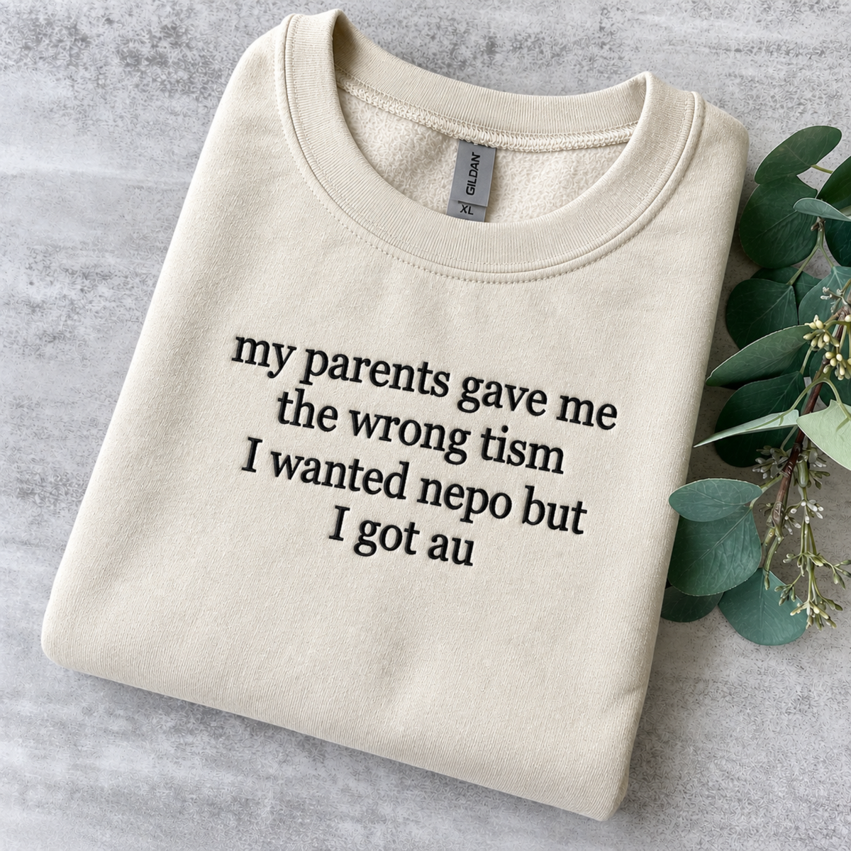A folded cream sweatshirt with black printed text reading 'my parents gave me the wrong tism I wanted nepo but I got au' on a gray textured surface, styled with green eucalyptus leaves on the right side.