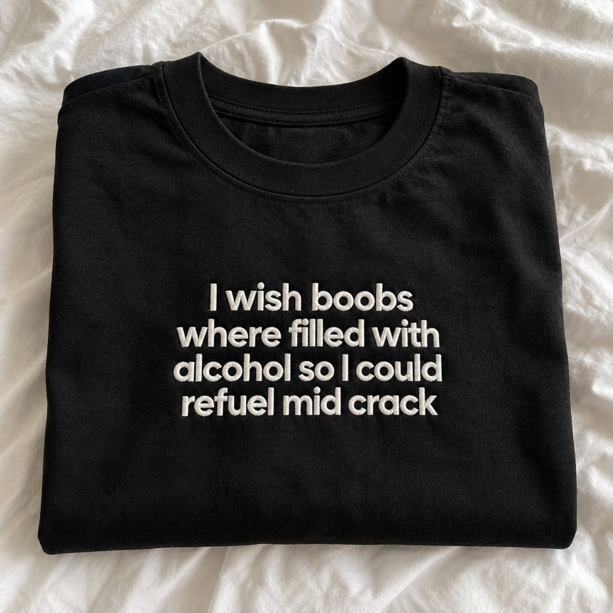 A folded black t-shirt with white text that reads 'I wish boobs where filled with alcohol so I could refuel mid crack', laid flat on a white wrinkled fabric background.