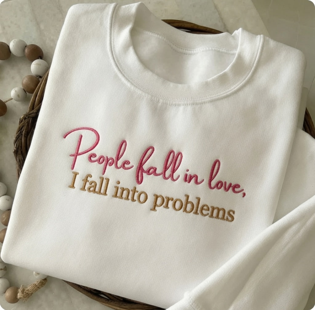 White crewneck sweatshirt neatly folded on a woven basket surface, embroidered with red cursive text reading "People fall in love," above tan block lettering reading "I fall into problems." A string of wooden beads is visible along the left side.