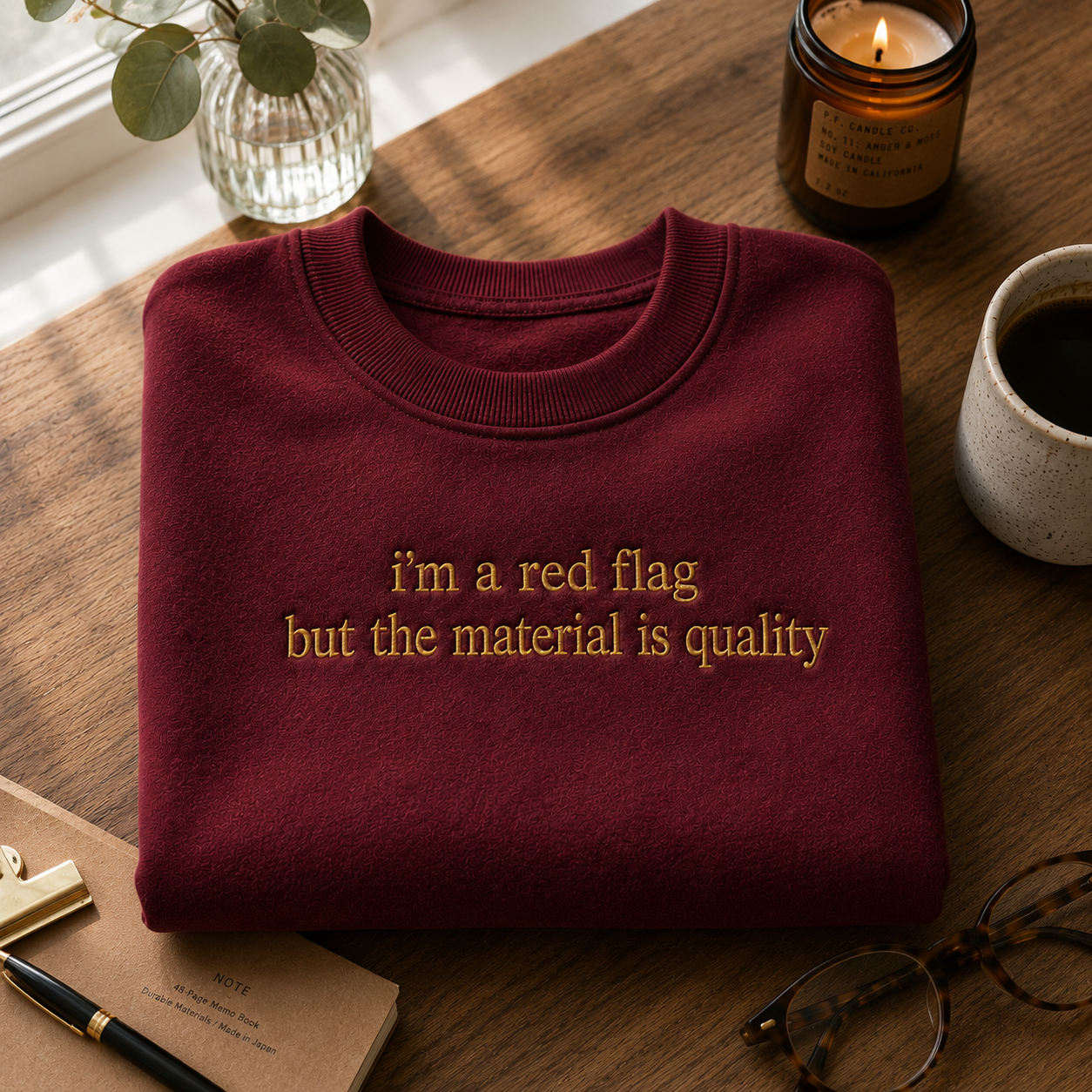 Maroon crewneck sweatshirt neatly folded on a wooden table next to a lit candle, glasses, and a notebook, featuring gold embroidered text that reads "i'm a red flag but the material is quality".