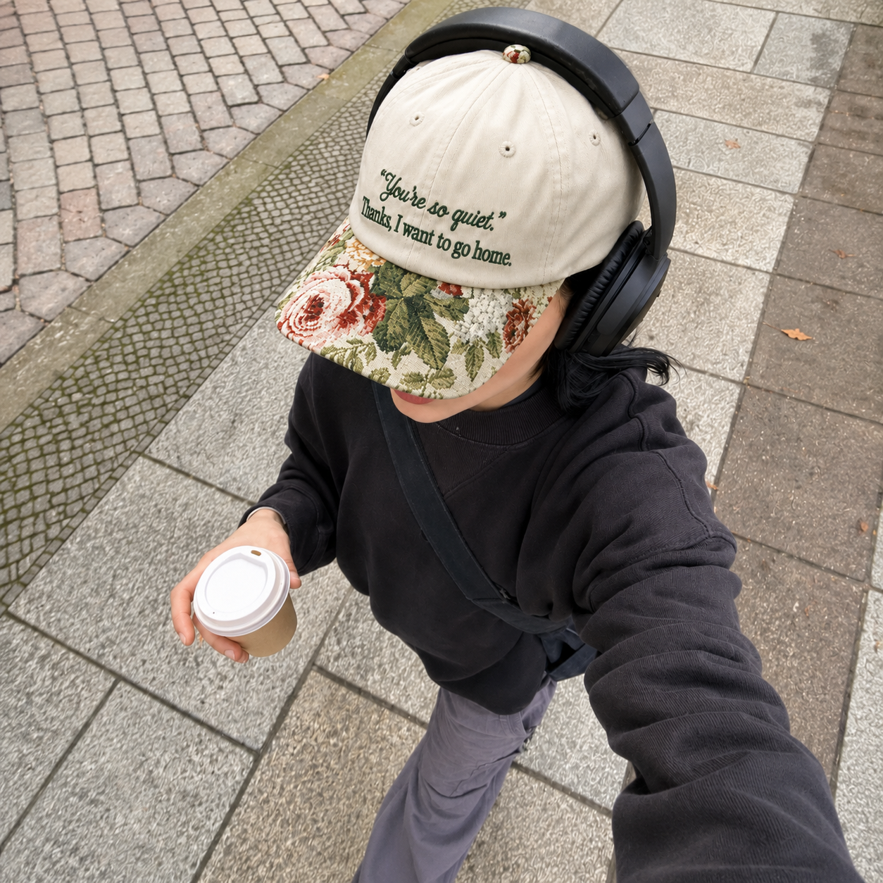High-angle view of a woman walking outdoors wearing a beige embroidered baseball cap with black text 'You're so quiet. Thanks, I want to go home.', a floral visor, over-ear headphones, and holding a coffee cup.