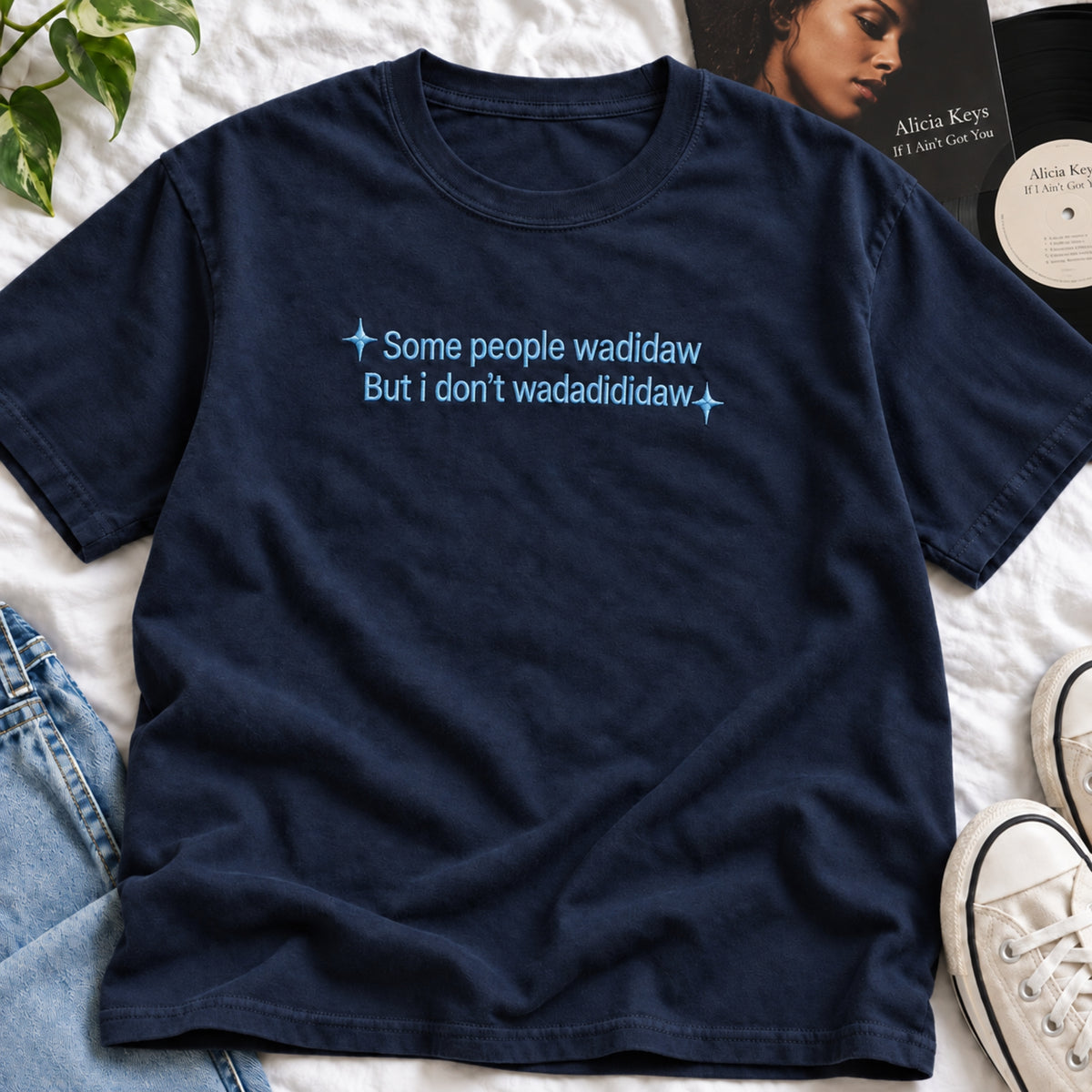 A navy blue short-sleeve t-shirt with light blue text reading 'Some people wadidaw But i don't wadadididaw' printed on the chest, styled in a flat-lay with jeans, sneakers, and a record sleeve on a white fabric background.