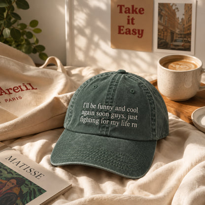 A dark teal-green baseball cap with white embroidered text reading 'I'll be funny and cool again soon guys, just fighting for my life rn' is styled on a beige bed with a canvas tote bag, a coffee cup, and a book in a warm, sunlit room.