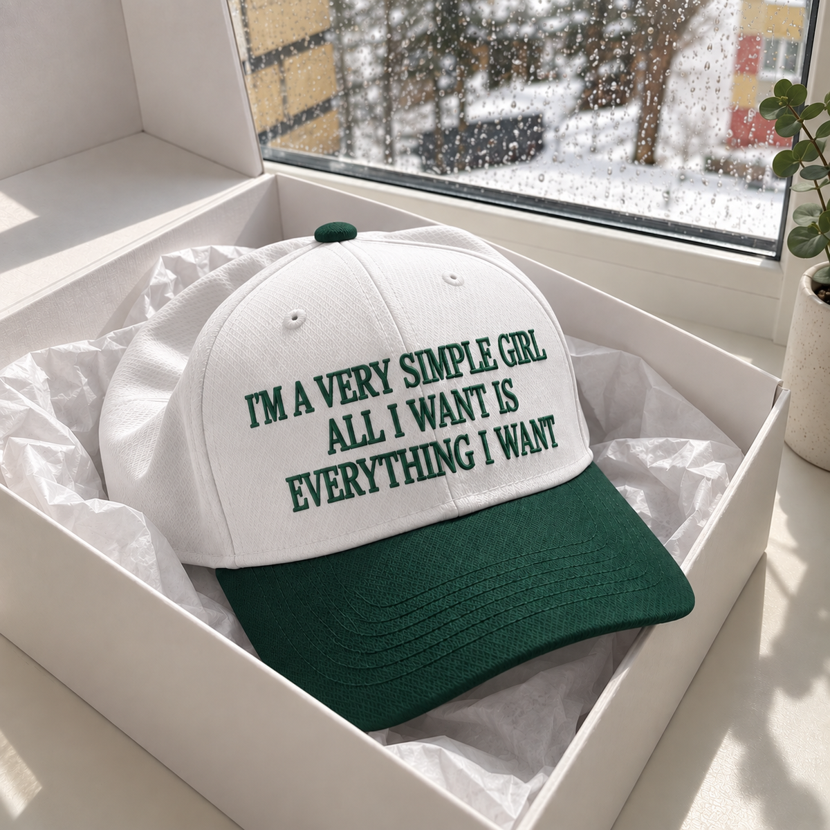 A white baseball cap with a dark green brim and green embroidered text reading 'I'M A VERY SIMPLE GIRL ALL I WANT IS EVERYTHING I WANT', displayed inside a white gift box with tissue paper. The box is set on a windowsill with a snowy outdoor view and a potted plant in the background.
