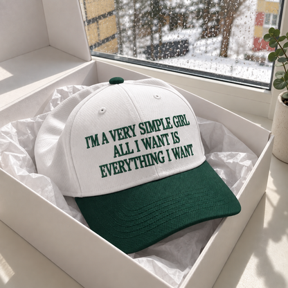 A white baseball cap with a dark green brim and green embroidered text reading 'I'M A VERY SIMPLE GIRL ALL I WANT IS EVERYTHING I WANT', displayed inside a white gift box with tissue paper. The box is set on a windowsill with a snowy outdoor view and a potted plant in the background.