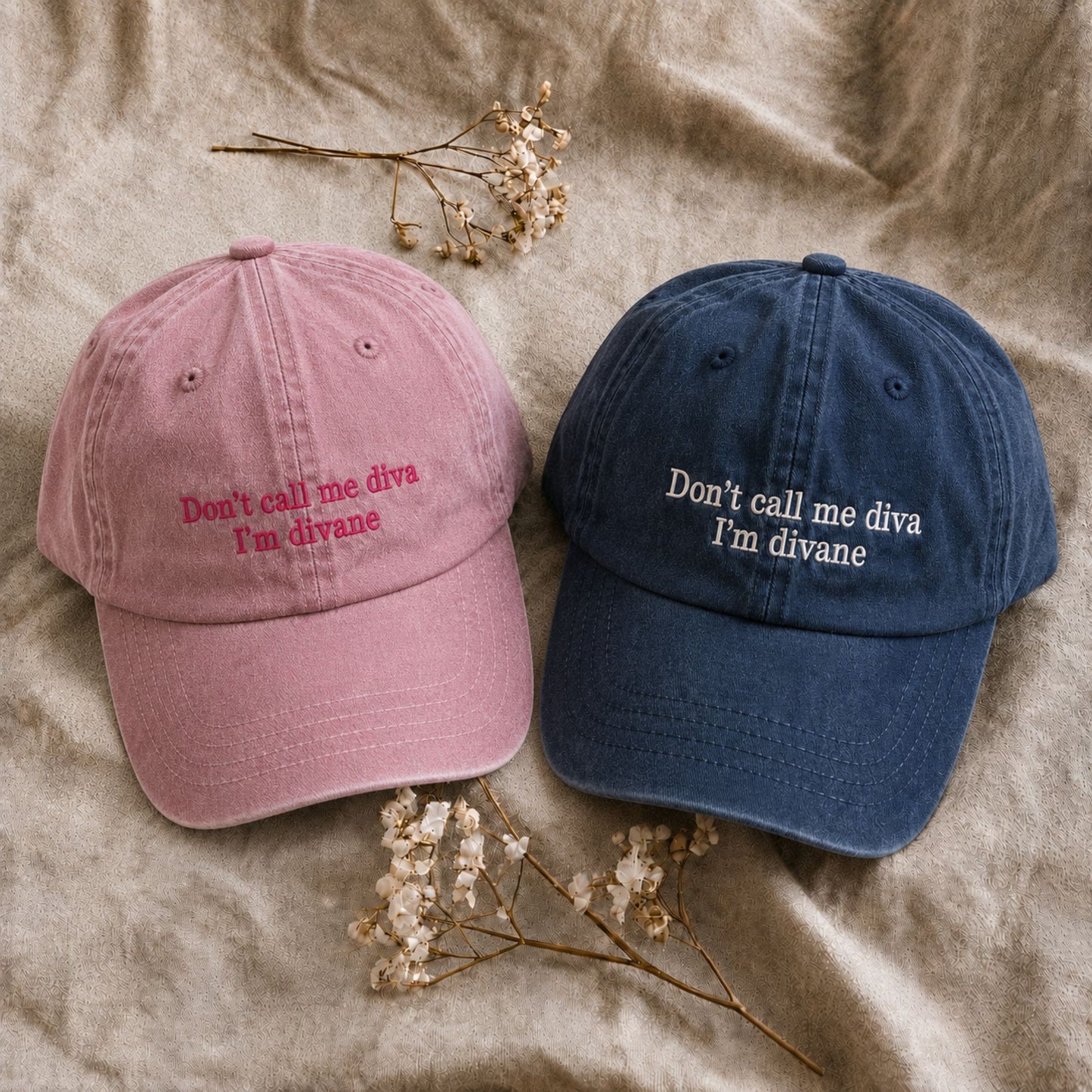 Two baseball caps laid flat on a beige fabric background with small dried flowers. One cap is dusty pink with pink embroidered text reading 'Don't call me diva I'm divane,' and the other is dark navy blue with white embroidered text reading 'Don't call me diva I'm divane.