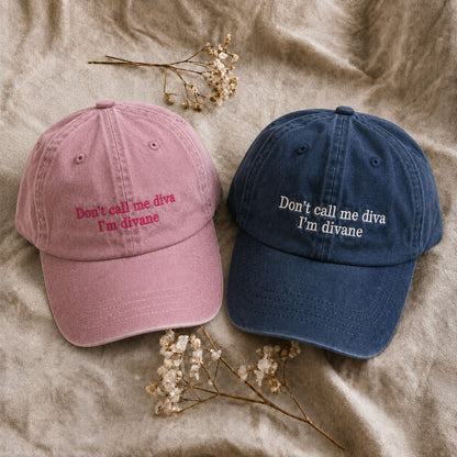 Two baseball caps laid flat on a beige fabric background with small dried flowers. One cap is dusty pink with pink embroidered text reading 'Don't call me diva I'm divane,' and the other is dark navy blue with white embroidered text reading 'Don't call me diva I'm divane.