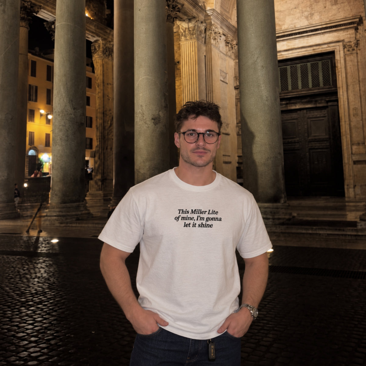 Male model wearing a white T-shirt with black embroidered text reading “This Miller Lite of mine, I’m gonna let it shine,” standing outdoors at night in front of large stone columns.