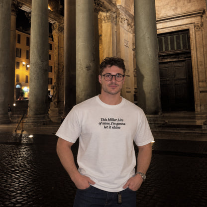 Male model wearing a white T-shirt with black embroidered text reading “This Miller Lite of mine, I’m gonna let it shine,” standing outdoors at night in front of large stone columns.